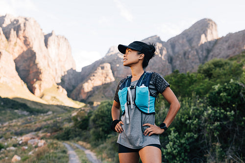 Trail runner in cap relaxing in valley. Sportswoman looking away and smiling preparing for hike.