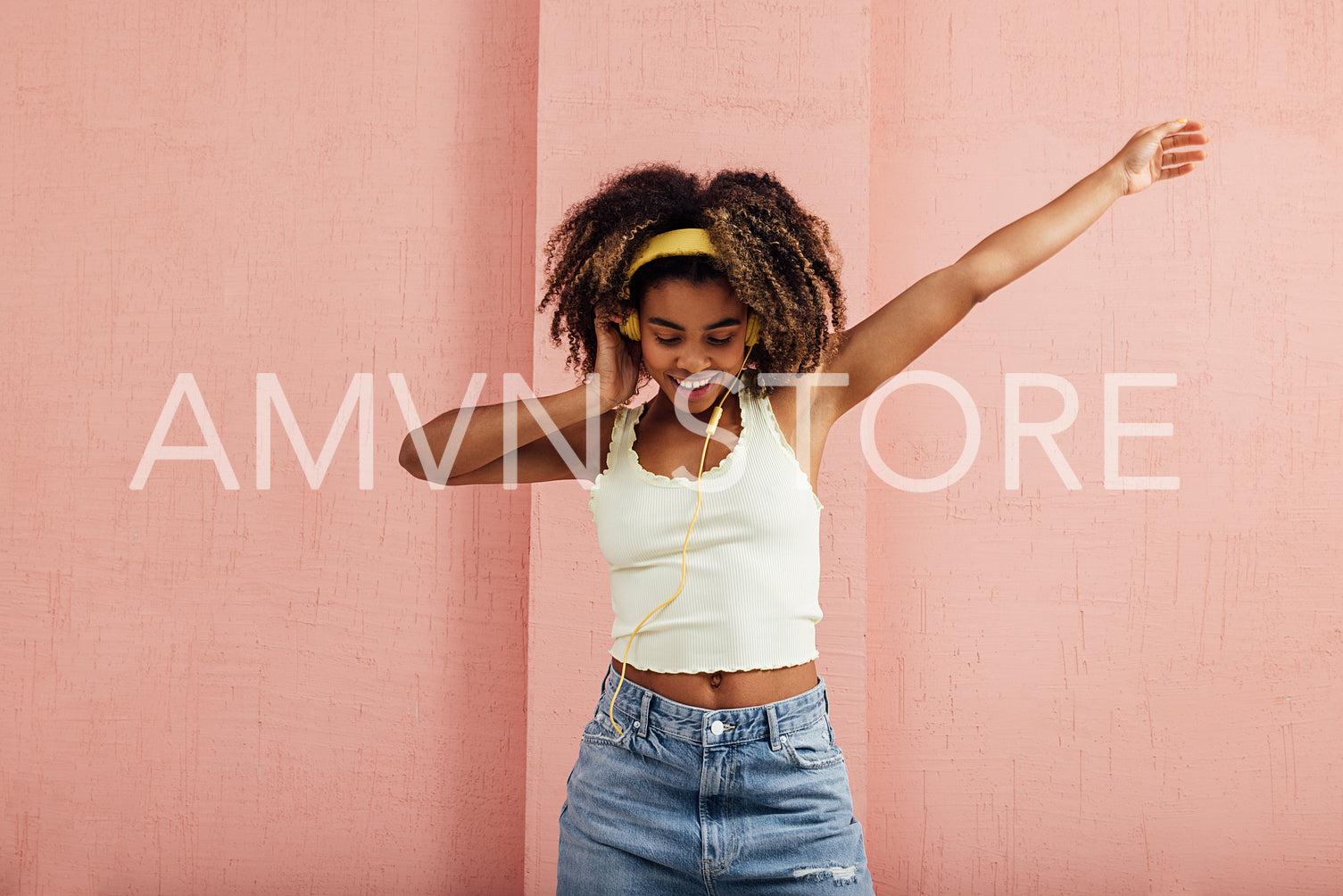 Cheerful woman with curly hair wearing yellow headphones dancing against a wall