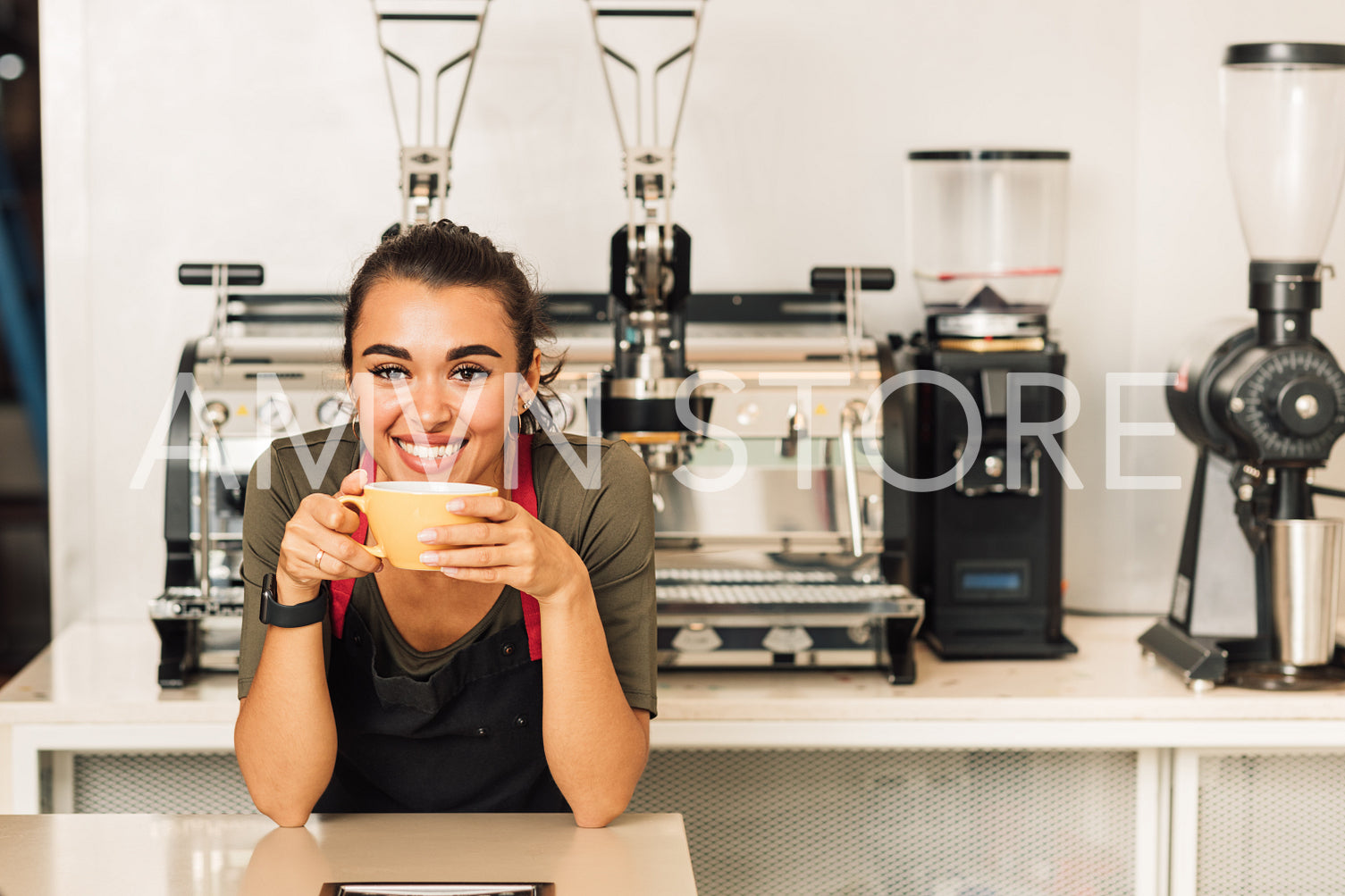 Smiling woman in apron leaning on a table while drinking coffee. Portrait of a waitress standing in front of the coffee machine.	