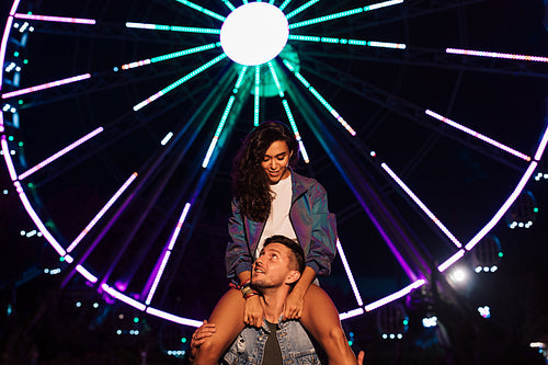 Smiling woman sitting on the shoulders of her boyfriend and looking at him. Young couple at night against ferris wheel with colorful lights.