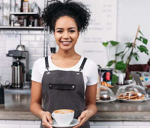 Smiling waitress with cup of coffee. African American woman working as a barista holding a cup with a freshly prepared cappuccino.
