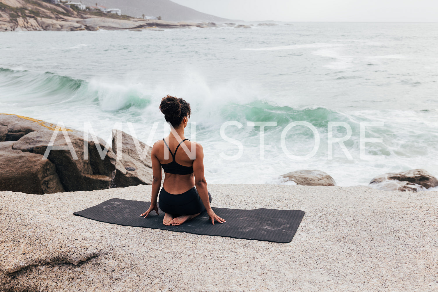 Rear view of young woman sitting in Vajrasana pose on mat outdoors. Woman meditating while looking at ocean.