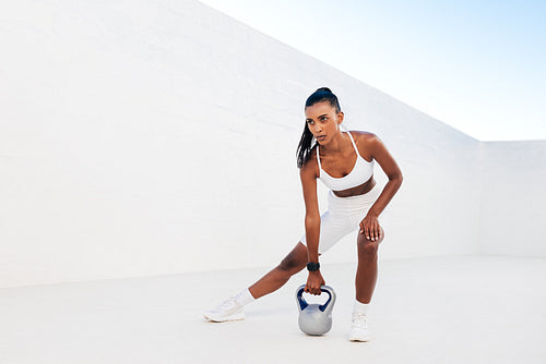 Full length of slim woman exercising with kettlebell outdoors. Professional fitness athlete doing intense training with weights in a white outdoor studio.