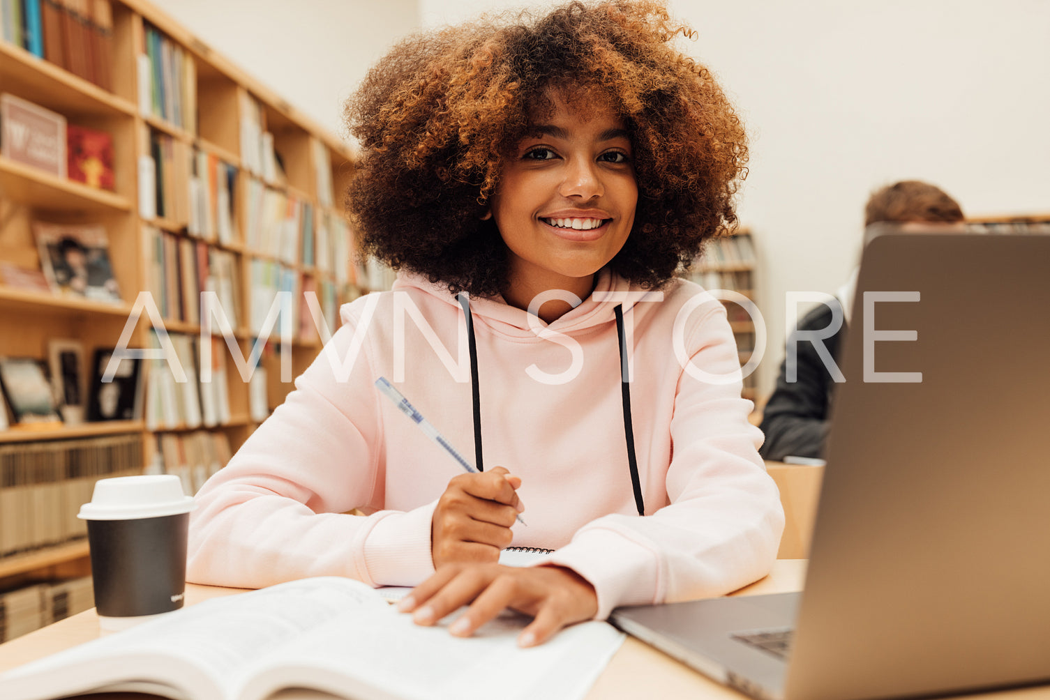 Portrait of smiling young student studying in library. Young female at library desk looking at camera.