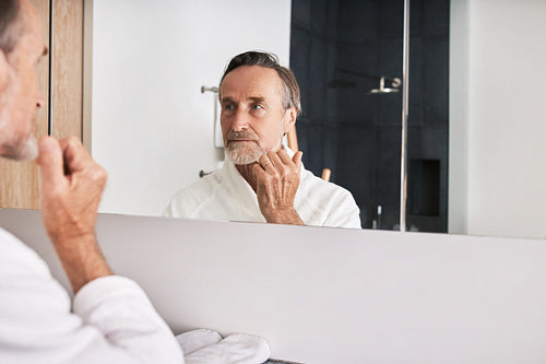 Handsome senior man wearing white bathrobe examine his skin in bathroom at morning