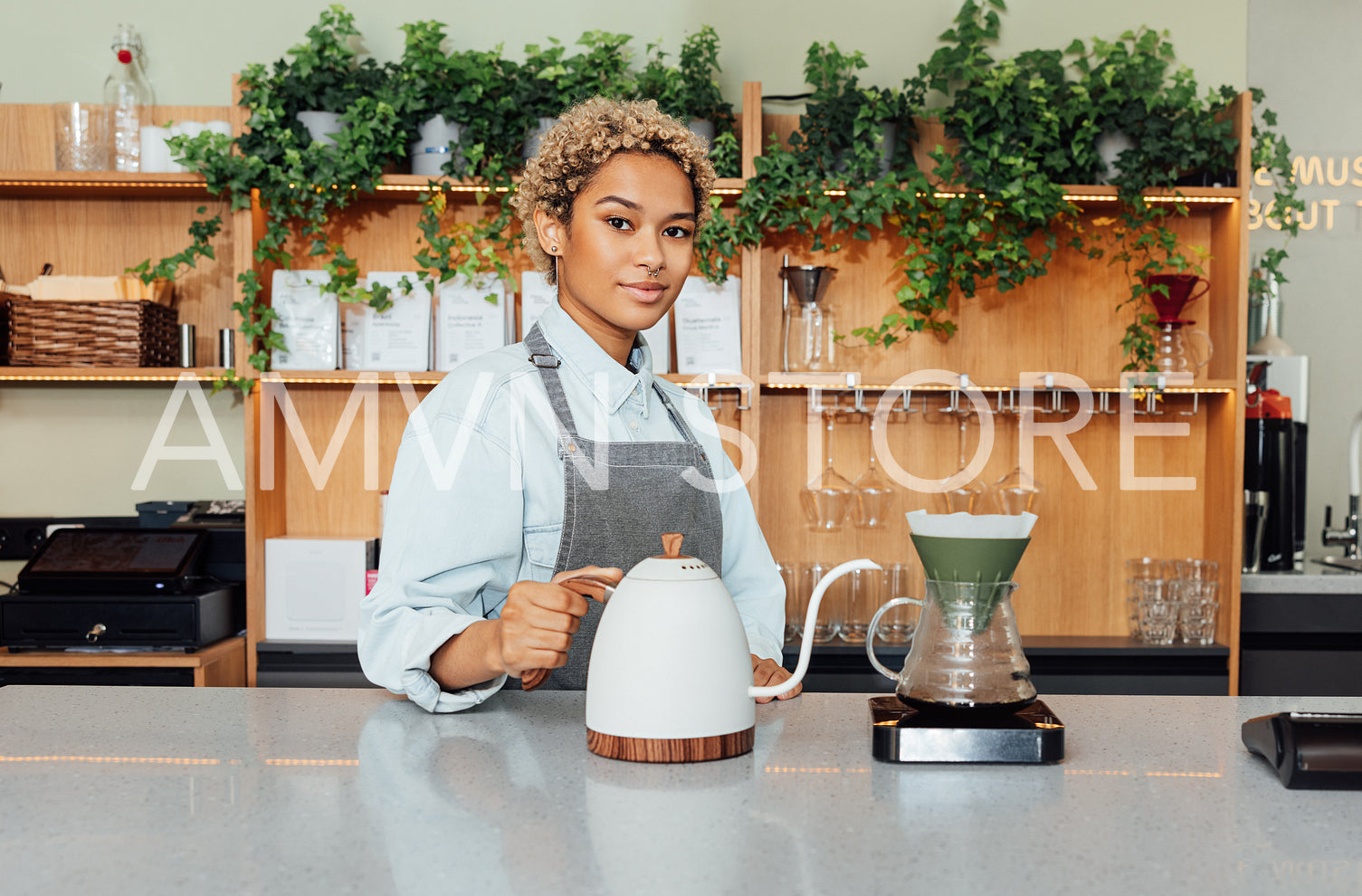 Portrait of a young barista at a counter in a cafe. Young woman working as a barista holding kettle looking at camera.