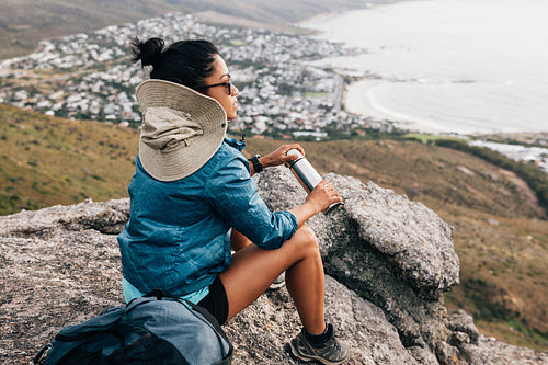 Rear view of a woman hiker in sports clothes opening a thermos while sitting on a cliff and looking at the view