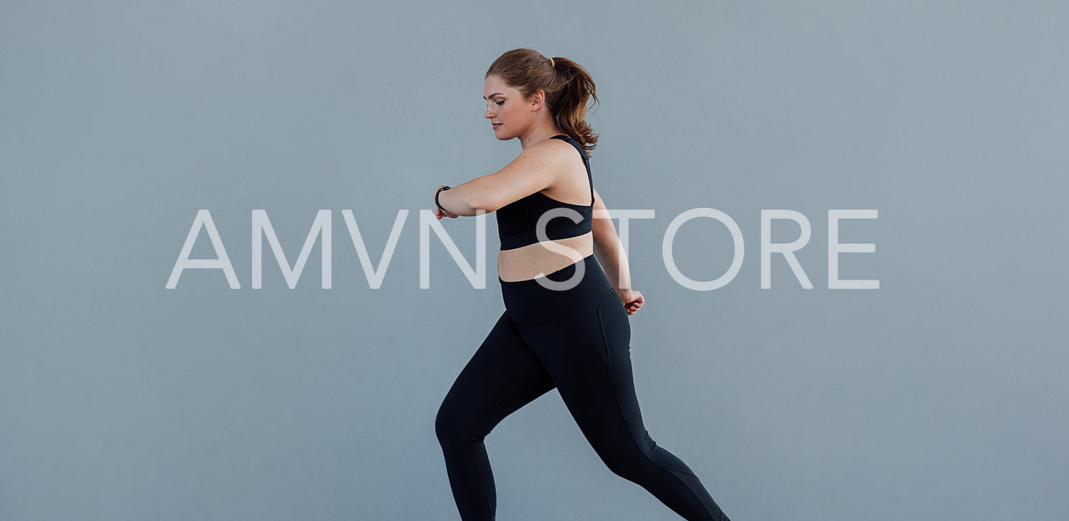 Plus-size female jogging at a grey wall. Woman with a plus-size body running outdoors and looking at her smartwatch.