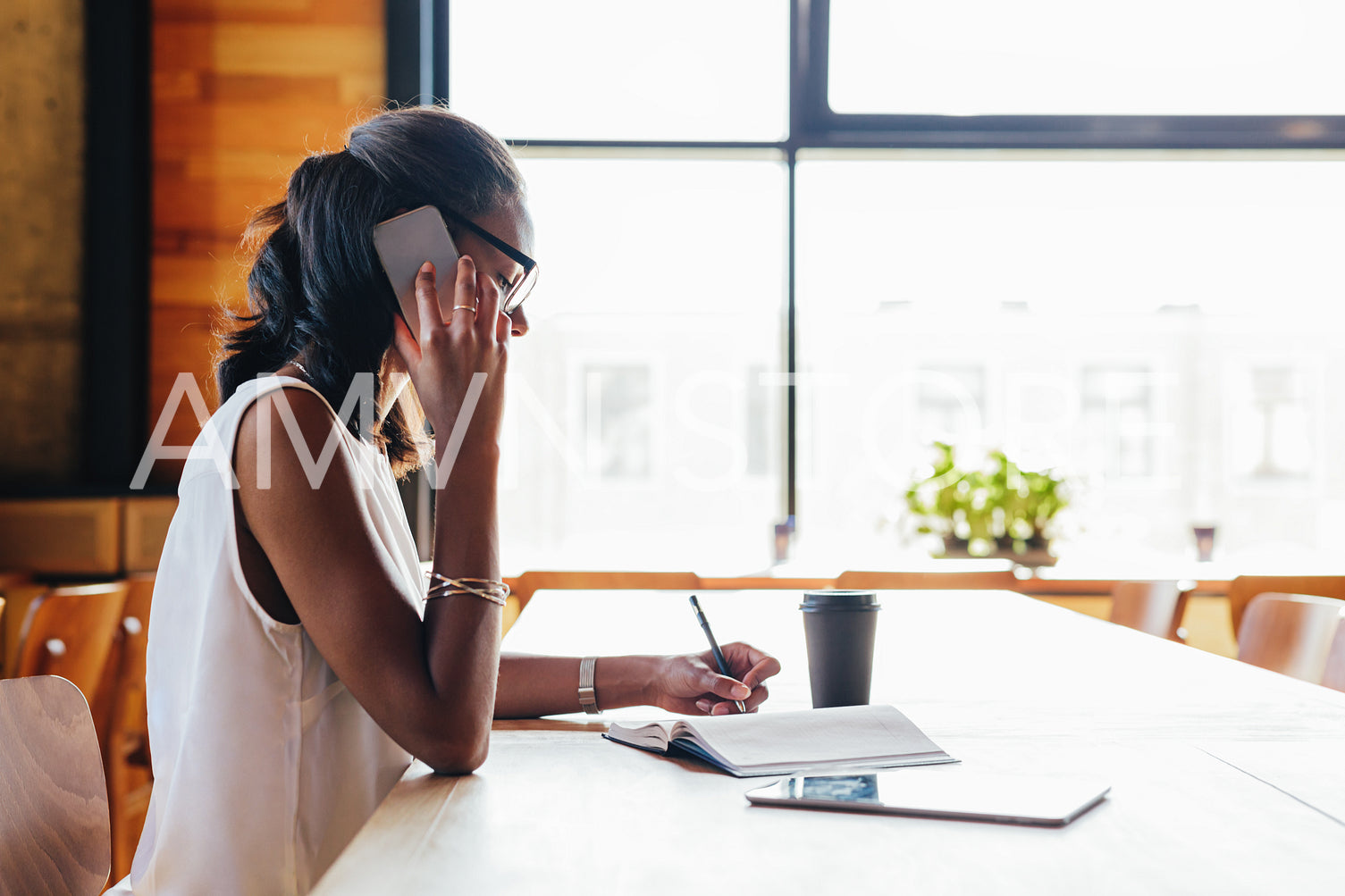 Businesswoman writing on notebook while talking on smartphone in cafe	