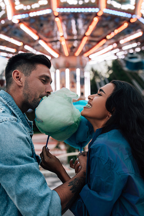 Young happy couple biting a cotton candy in the evening in an amusement park