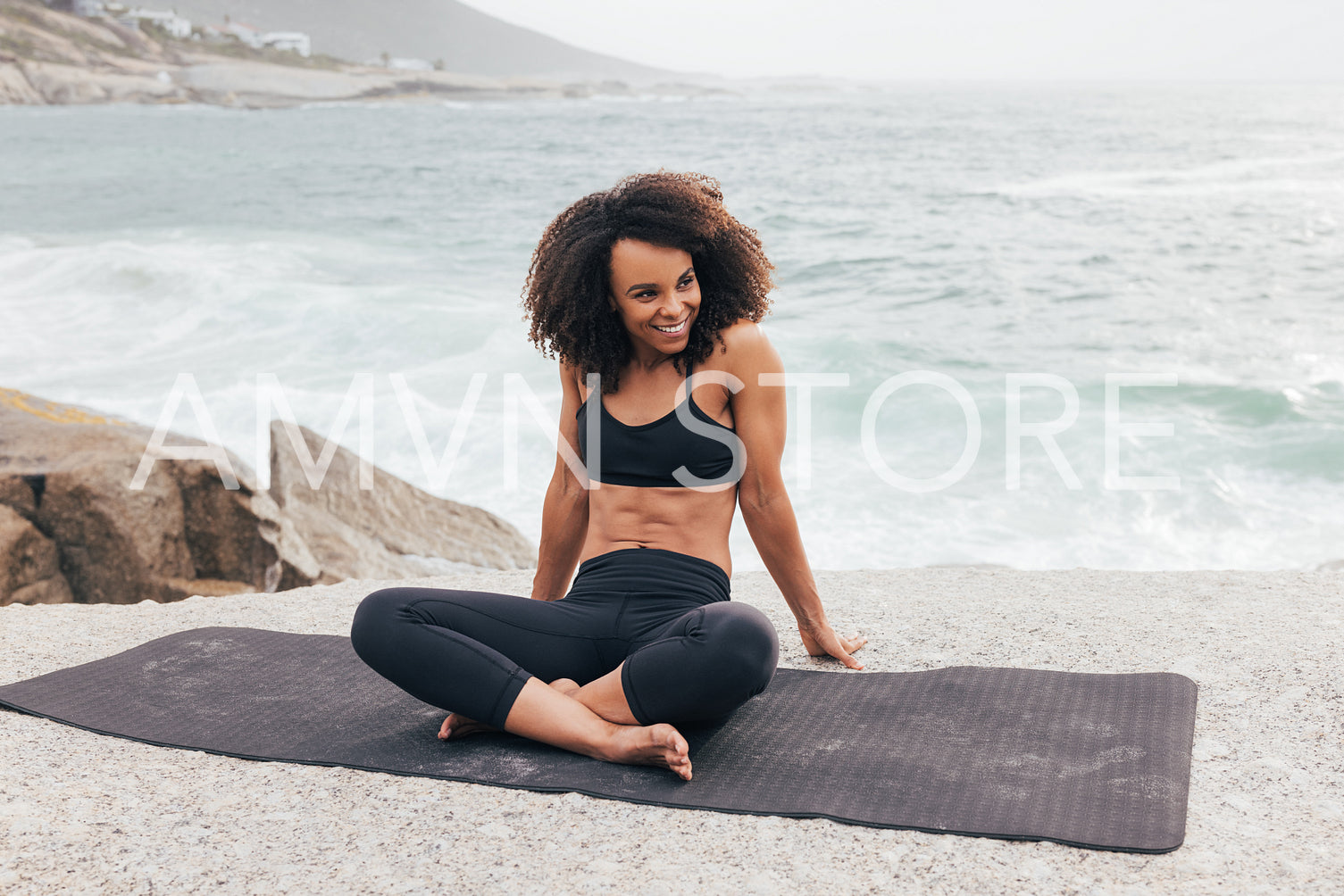 Smiling female relaxing on mat and looking away. Woman with crossed legs in sportswear by ocean.