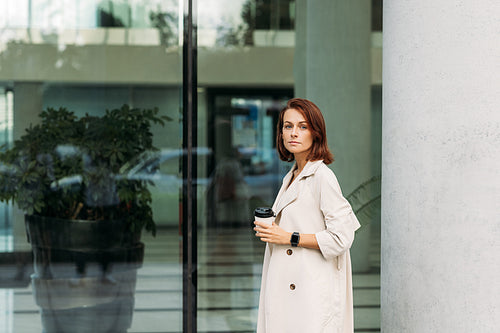 Stylish businesswoman in a coat holding a cup of coffee standing against a glass building