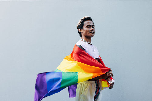 Side view of smiling male with closed eyes holding LGBT rainbow flag against grey wall