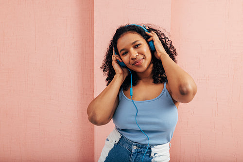 Plus size woman looking at camera wearing blue headphones against a pink wall