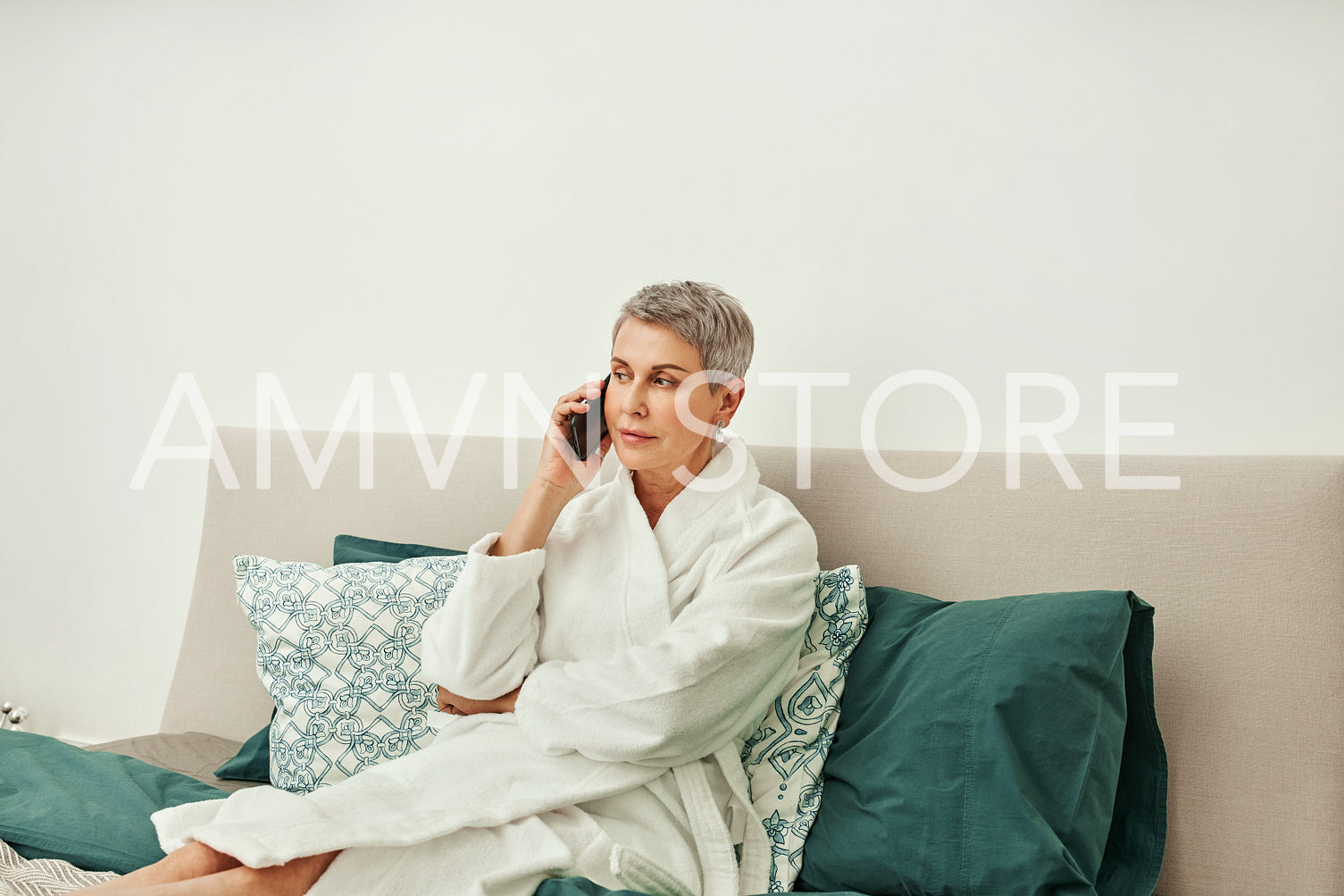 Mature woman in a bathrobe talking on mobile phone while lying on a bed in hotel room