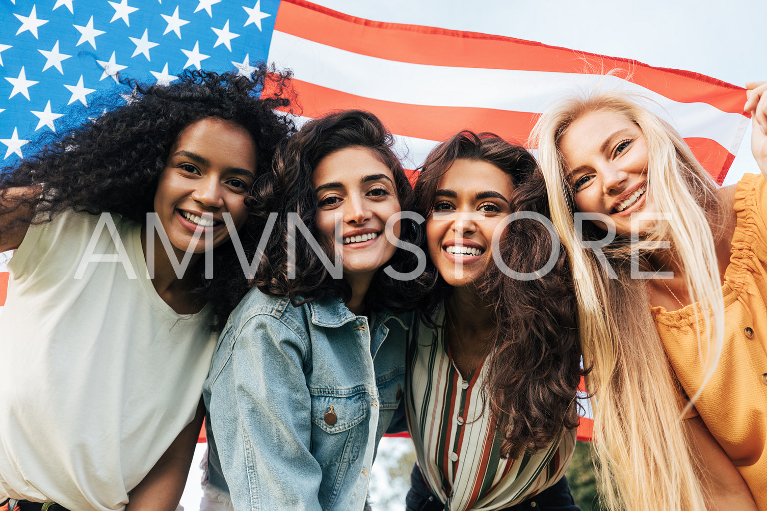 Four diverse women friends looking at the camera under the American flag. Cheerful females celebrating the 4th of July outdoors.