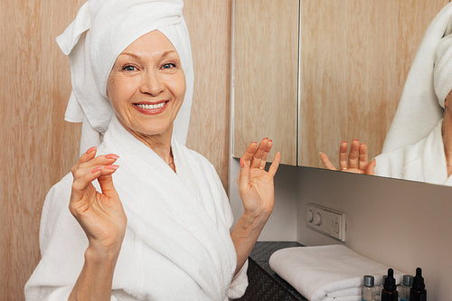 Smiling aged woman with a towel on her head looking at camera in a bathroom. Cheerful senior female in a bathrobe standing in a bathroom.