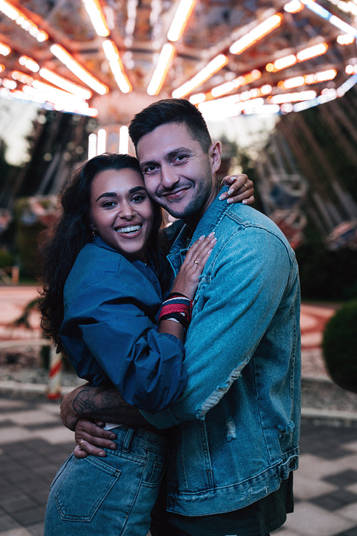 Portrait of a happy young couple in an amusement park at night. Boyfriend and girlfriend are looking at camera while standing against the carousel.