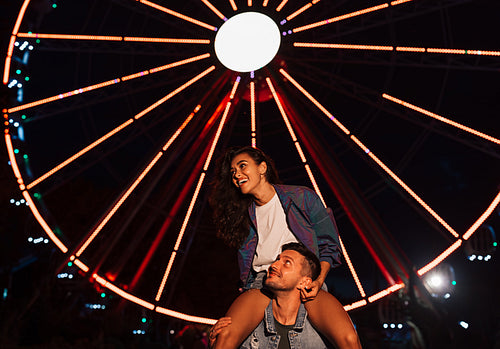 Young laughing female sitting on the shoulders of her boyfriend and looking away at night in an amusement park