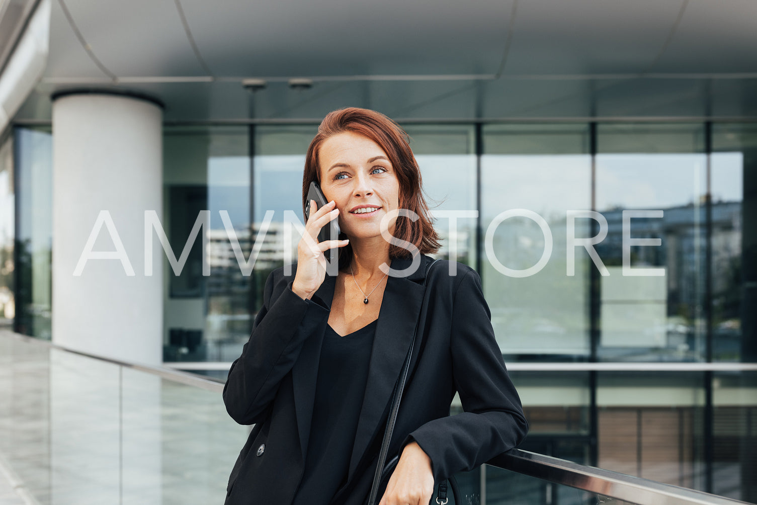 Smiling middle-aged business woman with ginger hair leaning on a railing talking on a mobile phone