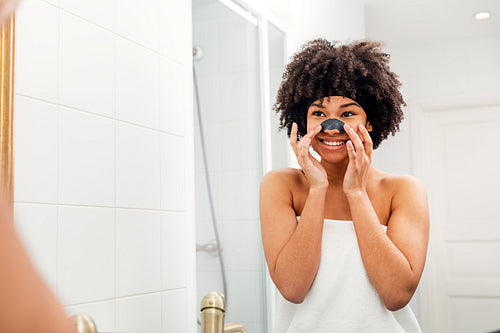 Smiling woman applying nose patch. Young female wrapped in white towel in bathroom.