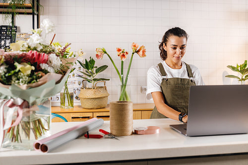 Woman wearing apron working in her flower shop. Young businesswoman typing on laptop indoors.