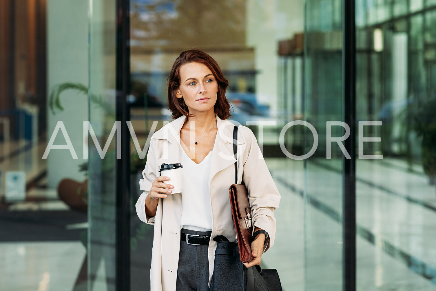 Middle-aged businesswoman holding coffee going out from the business building. Confident businesswoman with ginger hair at business building.