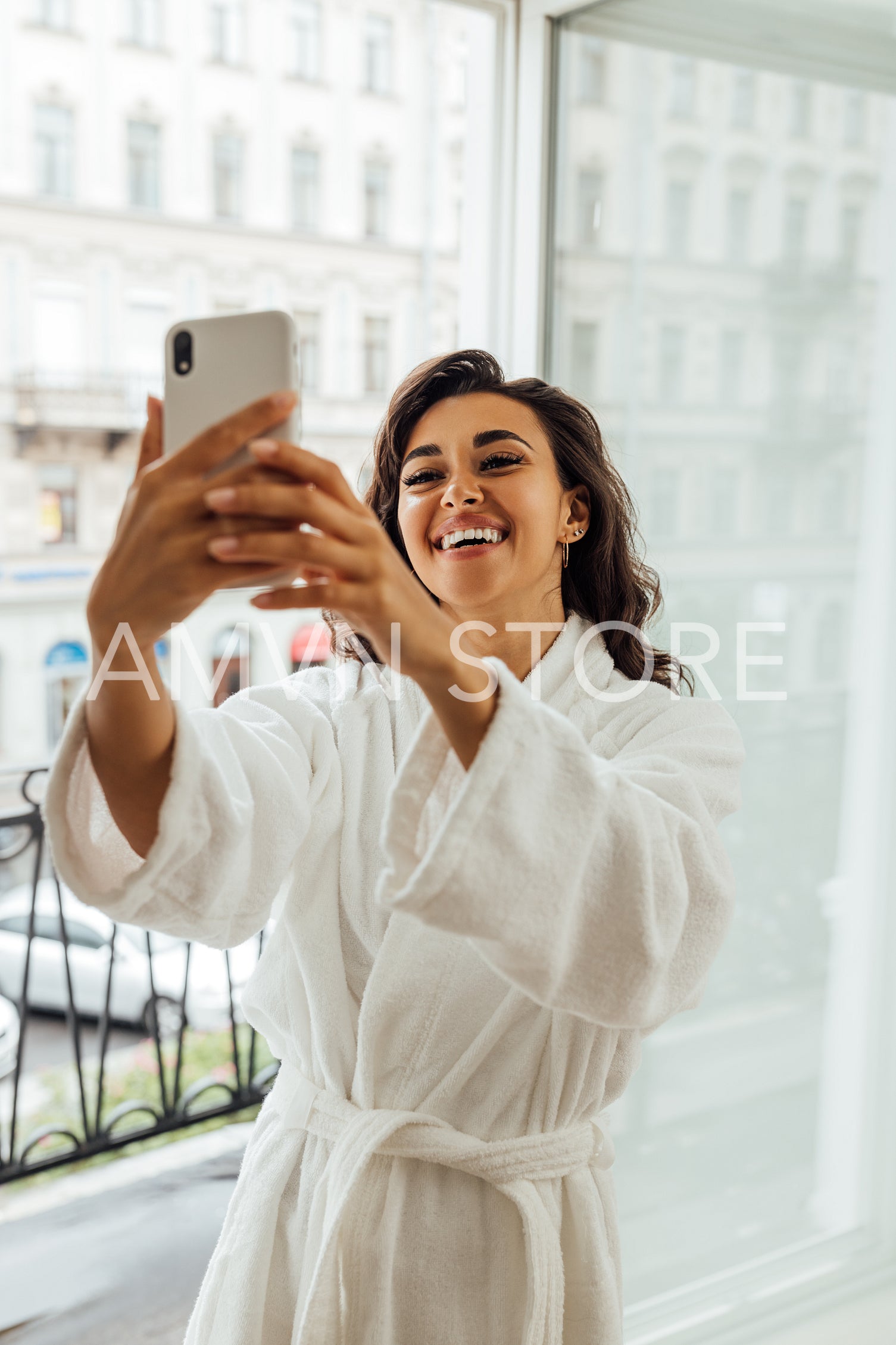 Happy woman in bathrobe taking selfie on smartphone. Smiling brunette live streaming from a balcony.	