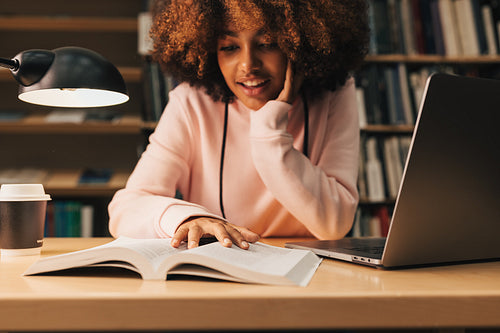 Smiling girl preparing exams in library at evening. Student sitting at desk and reading.