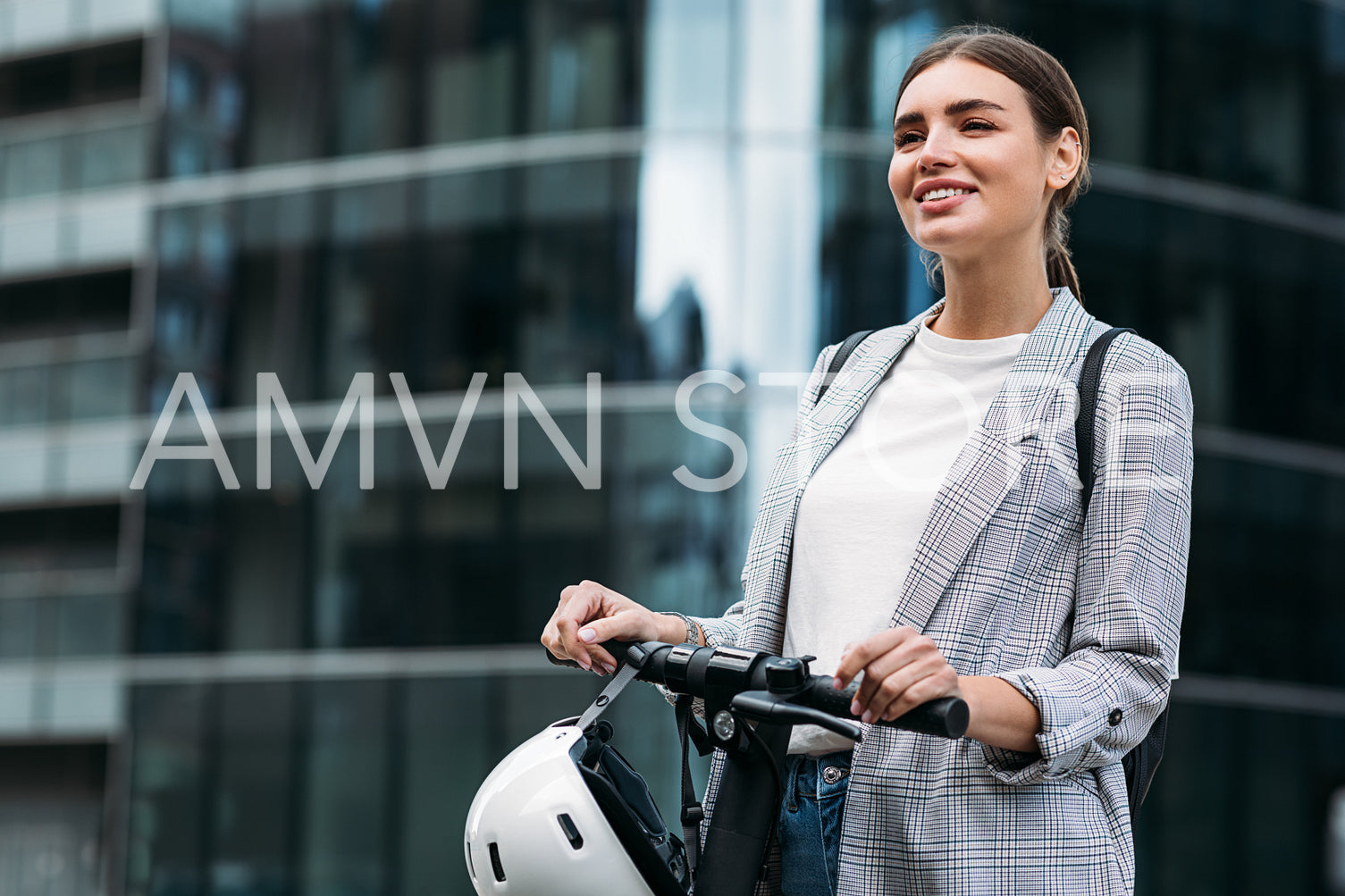 Smiling woman holding a handlebar of electric push scooter while standing at glass building