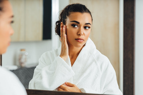 Young woman applying face cream in front of a mirror. Middle east female using a moisturizer after bath.