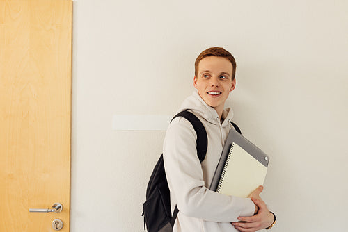Smiling university student with backpack and textbooks leaning wall