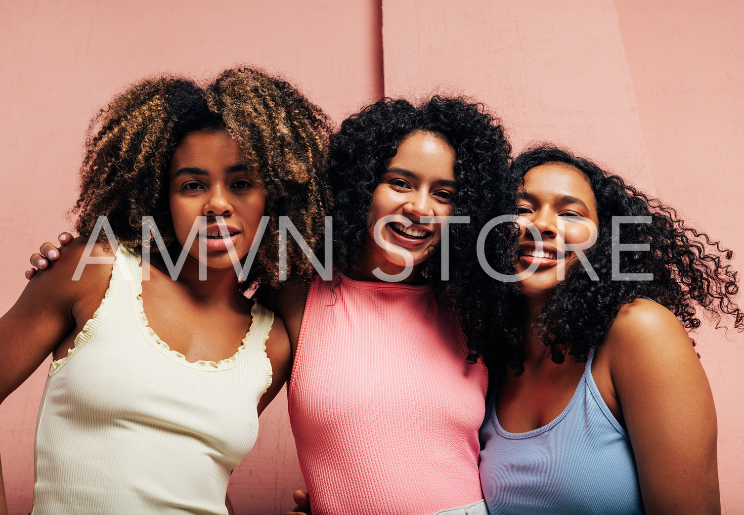 Three girlfriends in bright casual hugging each other looking at camera against a pink wall