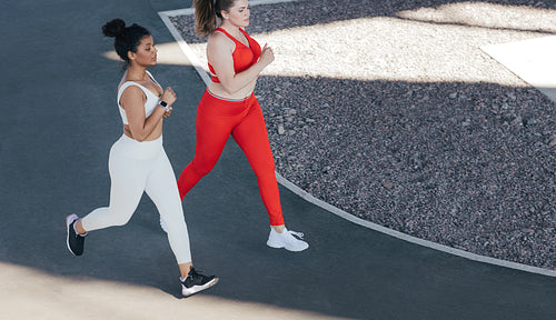Shot from above of two females running together outdoors. High angle of two female friends jogging together wearing sportswear in different colors.
