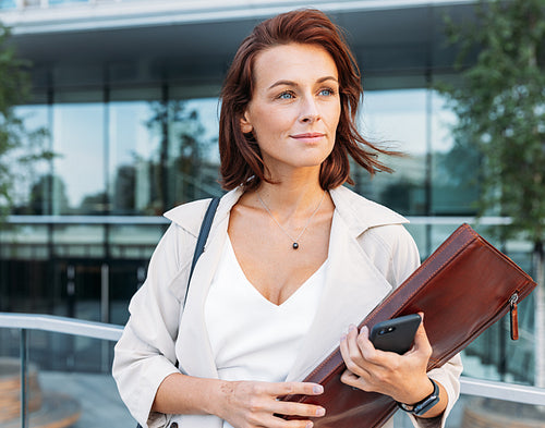 Portrait of a middle-aged attorney. Businesswoman with ginger hair holding a leather folder standing outdoors looking away.