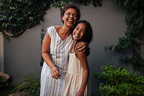 Portrait of granny and granddaughter embracing and laughing. Happy grandmother and kid standing together in backyard.