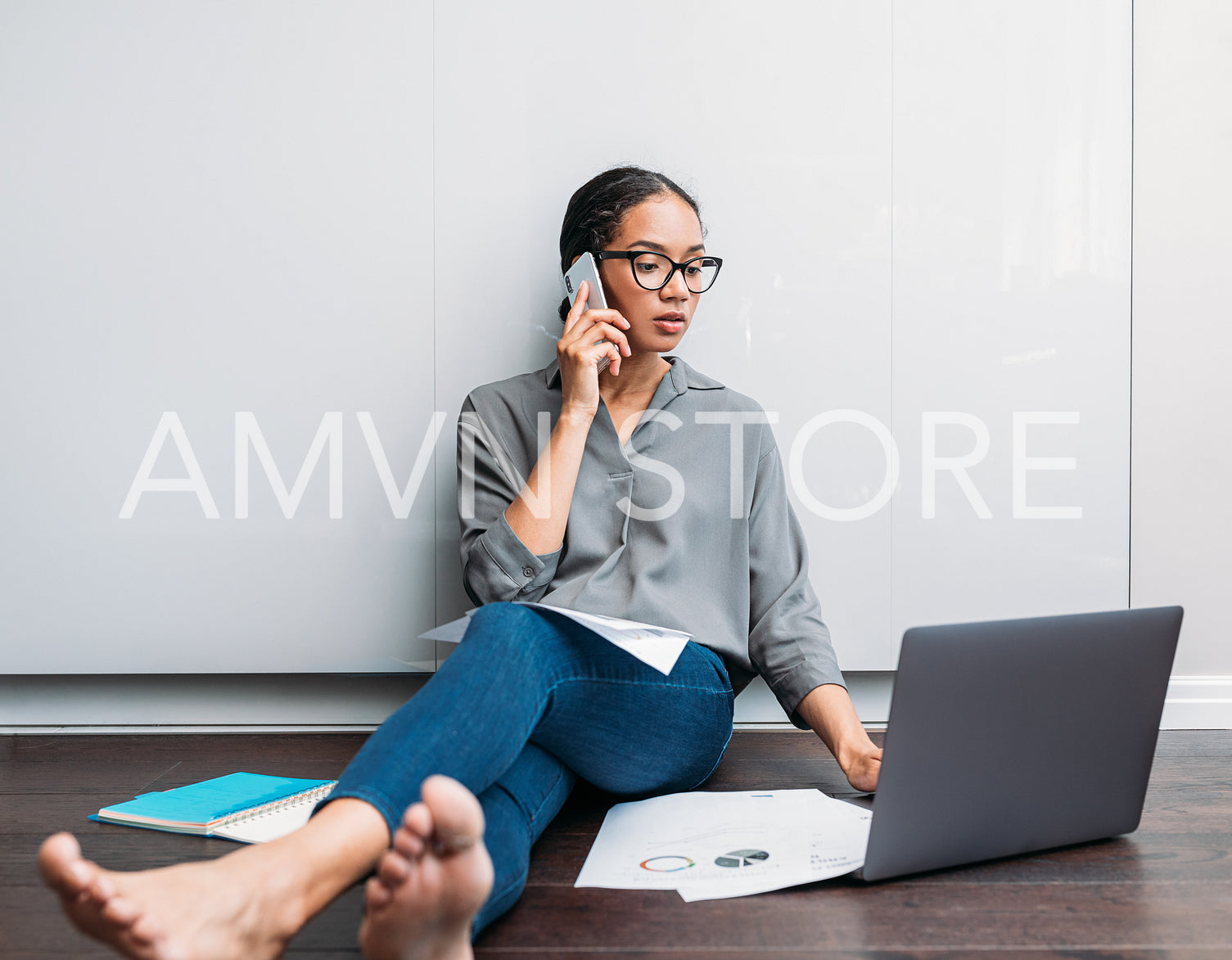 Young female using a laptop and talking on a cell phone while sitting on the floor at home