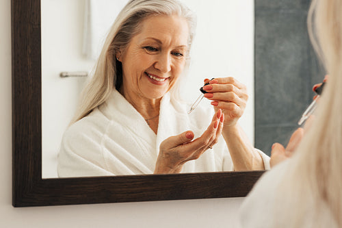 Reflection in mirror of a smiling aged woman with a pipette. Smiling senior female in a bathroom using liquid face treatment.