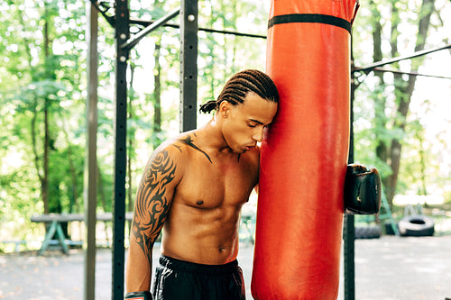 Exhausted kickboxer leaning on a punching bag outdoors. Young boxer resting during a workout on the sports ground.