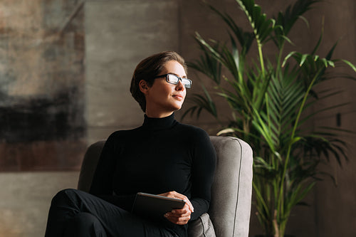 Young woman in black formal wear wearing glasses holding a digital tablet while sitting in her loft