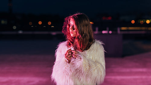 Young blonde woman standing on a roof in front of a pink neon light and looking away