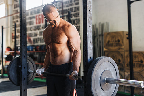 Side view of male athlete looking at his muscles while lifting barbell in gym