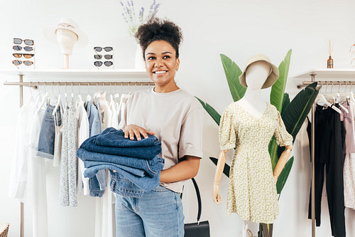 Smiling saleswoman standing in a small local store with a pile of jeans and looking away