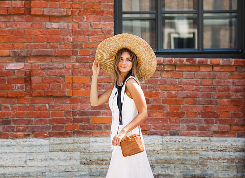 Young happy woman holding her big hat while walking on a street and looking back