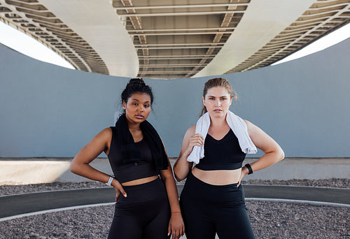 Portrait of two women in black fitness wear with towels on their necks. Young sportswomen are relaxing outdoors.