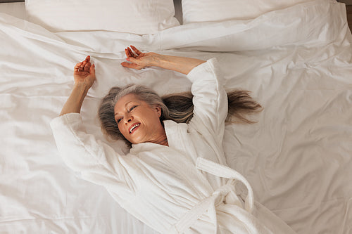 Smiling senior woman in a bathrobe lying on a bed and looking away. Female relaxing in a hotel room.