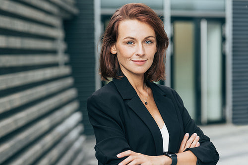 Confident middle-aged woman with crossed arms looking at the camera. Female in black formal clothes posing outdoors.