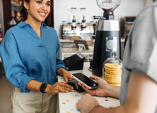 Hand of a customer at a cafe paying his bill using a mobile phone. Female entrepreneur holding a POS terminal while client paying using NFC technology.