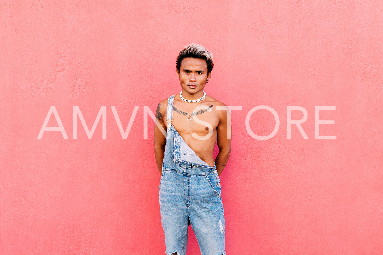 Portrait of a young handsome guy wearing jeans overalls standing against pink wall