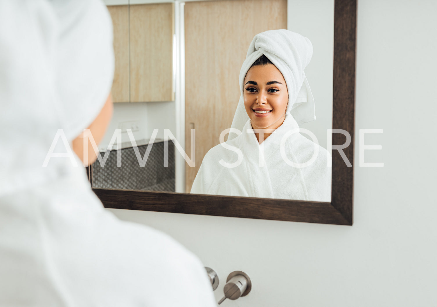 Woman in bathrobe looking at her reflection. Beautiful brunette with a white towel on her head.	